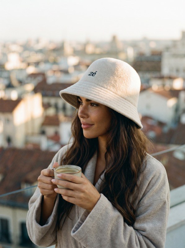 Woman wearing the zajé Oat hat with a robe and coffee on a rooftop at golden hour